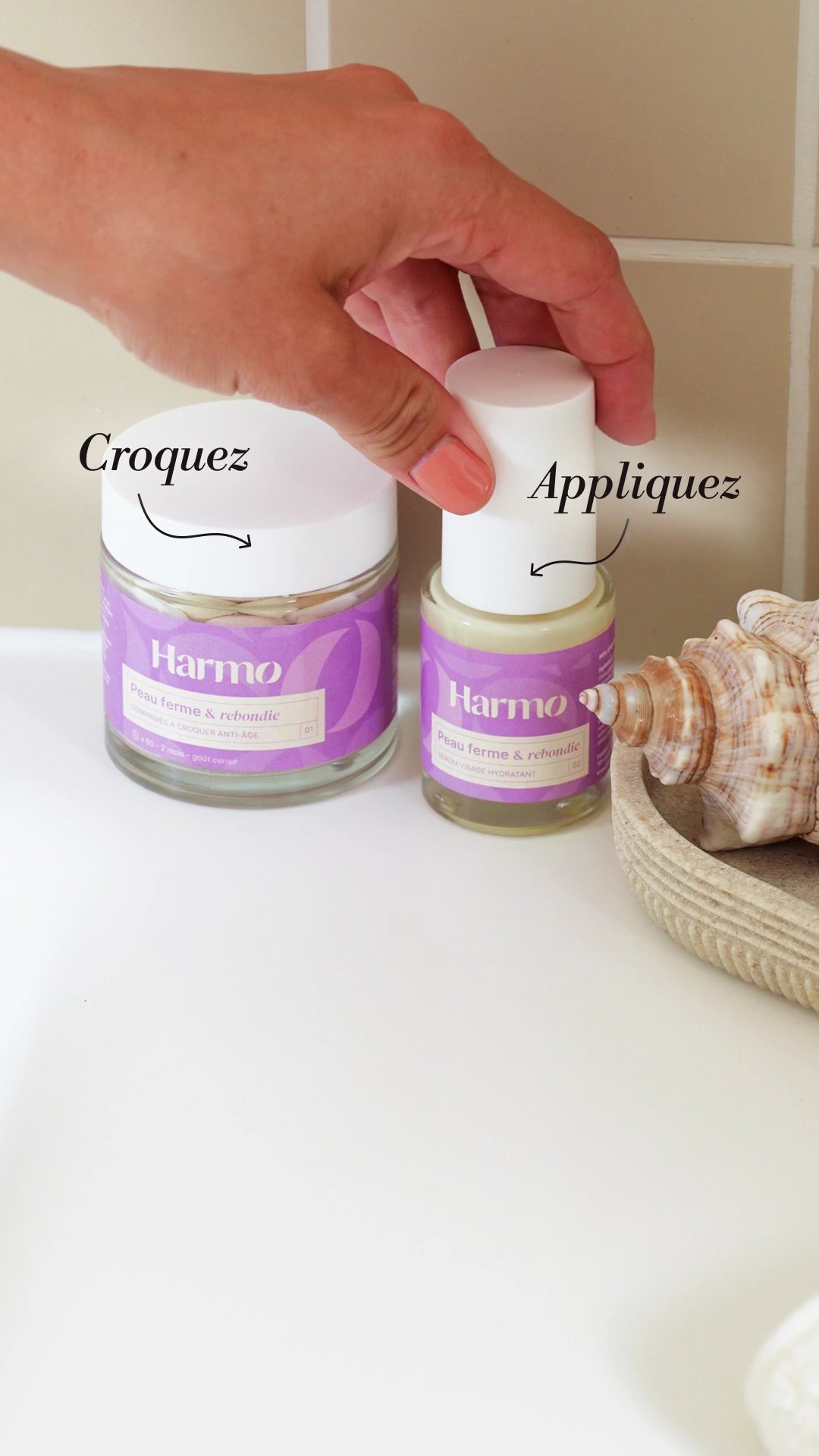 Une main qui attrape le sérum anti-âge du duo Harmo Skincare In&Out qui est à côté d'un pot de comprimés à croquer. 